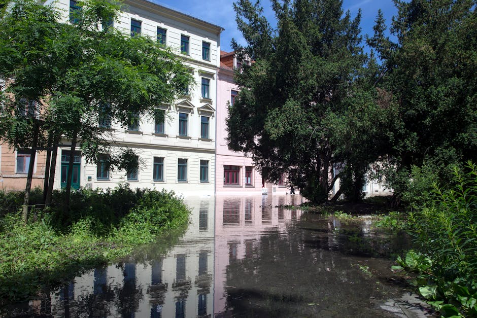 Photo of a flooded street on Uxbridge High Street with water extending across the road's surface, partially submerging the pavement and curbs. Several multi-storey residential buildings with white and