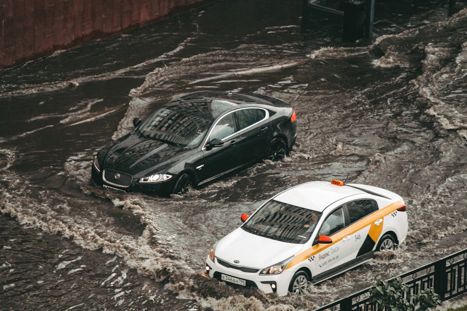 Photo of a flooded street on Uxbridge High Street with water extending across the road's surface, partially submerging the pavement and curbs. Several multi-storey residential buildings with white and