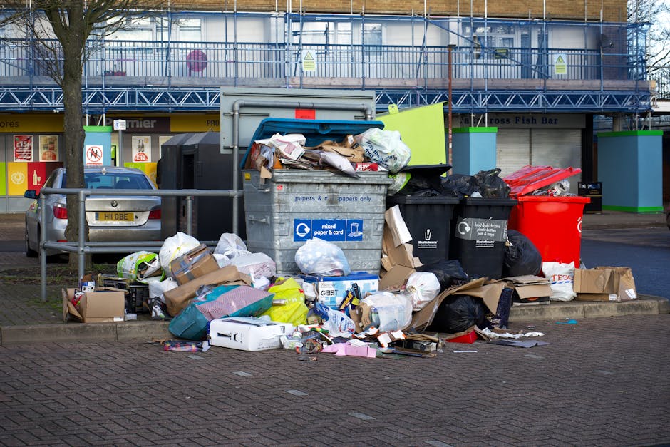 A large outdoor waste disposal area filled with various overflowing rubbish bins and scattered trash including cardboard boxes, paper, plastic bags, and discarded packaging on a paved sidewalk. Behind