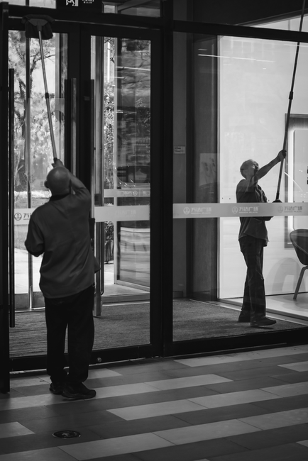 A professional cleaner from uxbridgecarpetcleaners.com is performing surface cleaning in a modern, well-lit living room. The cleaner, dressed in black attire with an apron, uses a broom to sweep the l